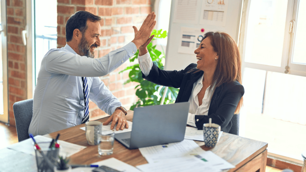 Two professionals in business attire celebrate a shared win with a high-five across a wooden desk in a bright modern office; a laptop and notebooks sit between them.