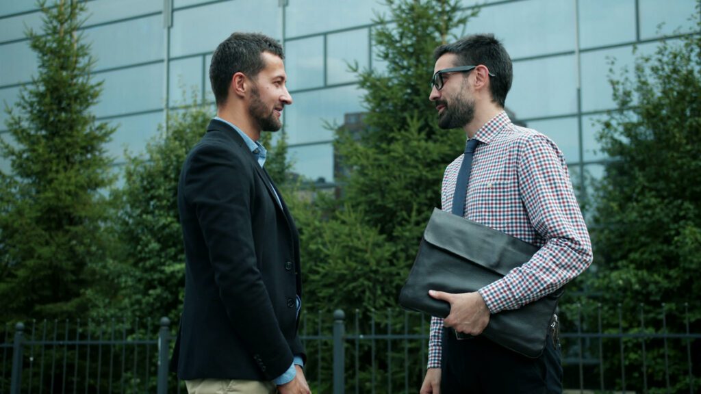 Two business professionals speak outside a modern civic-style building, suggesting a serious discussion about institutional leadership and public infrastructure.