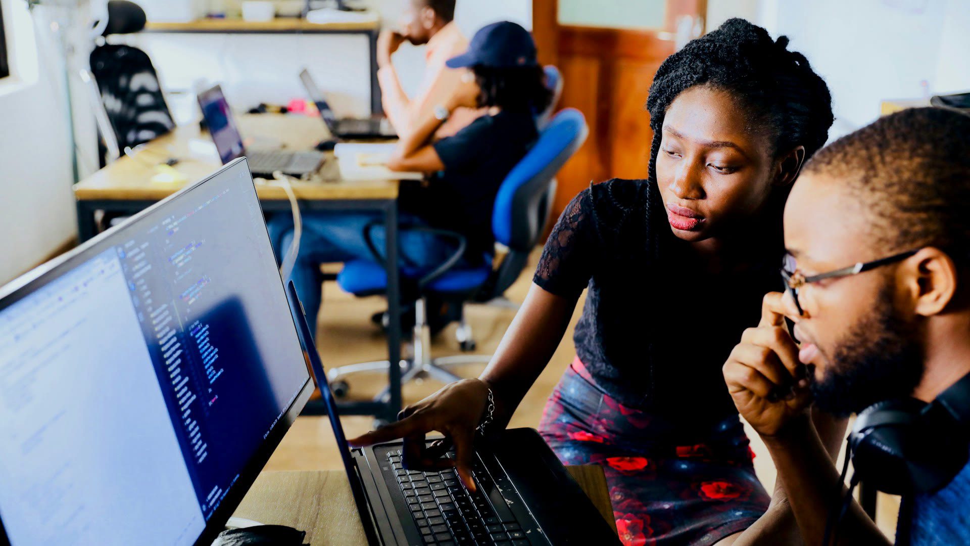 Two professionals collaborate at a bright modern office workstation, reviewing a large monitor together in a busy corporate workspace.