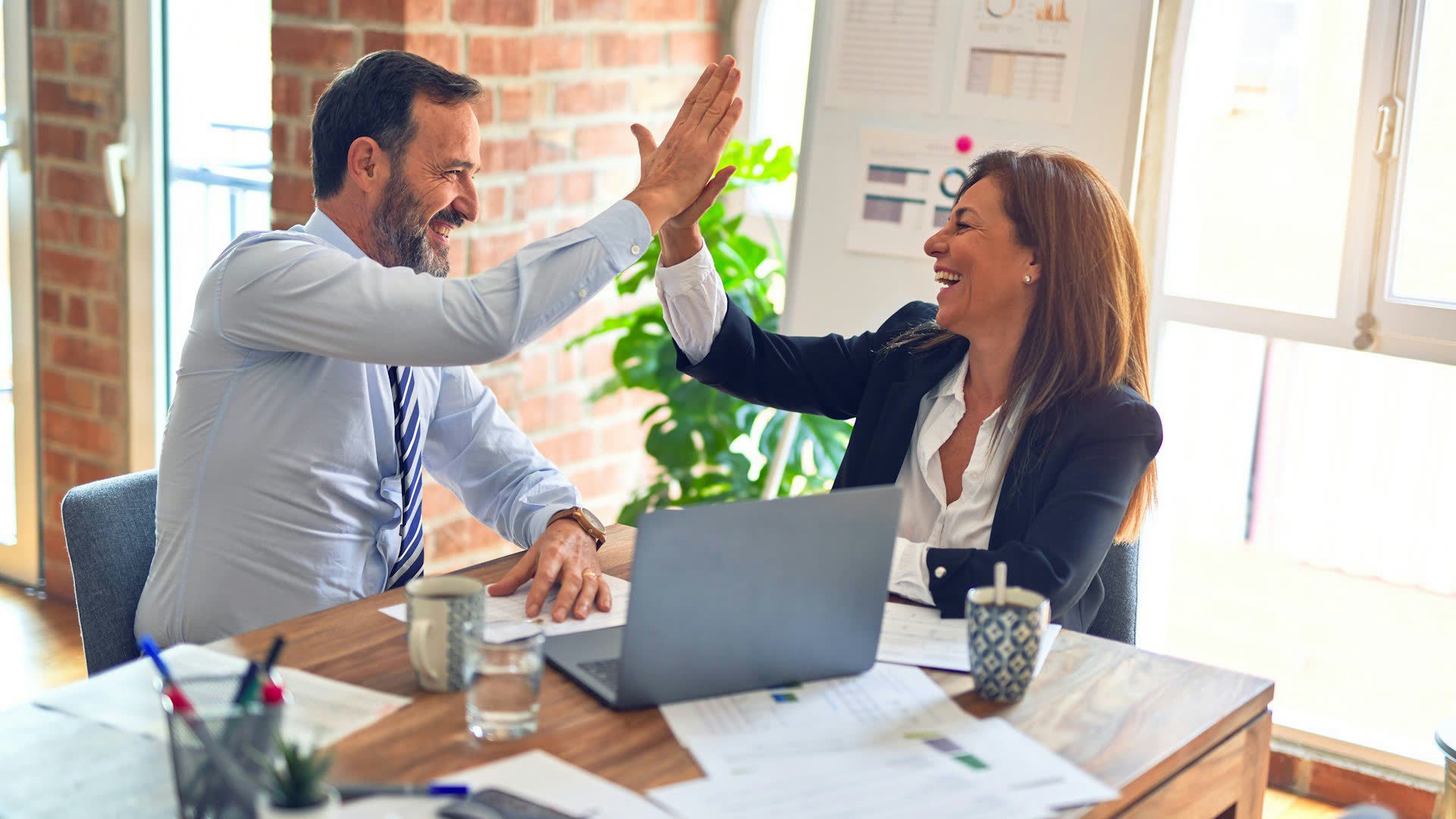 Two professionals high-five across a wooden desk in a bright modern office, smiling over an open laptop and work papers.
