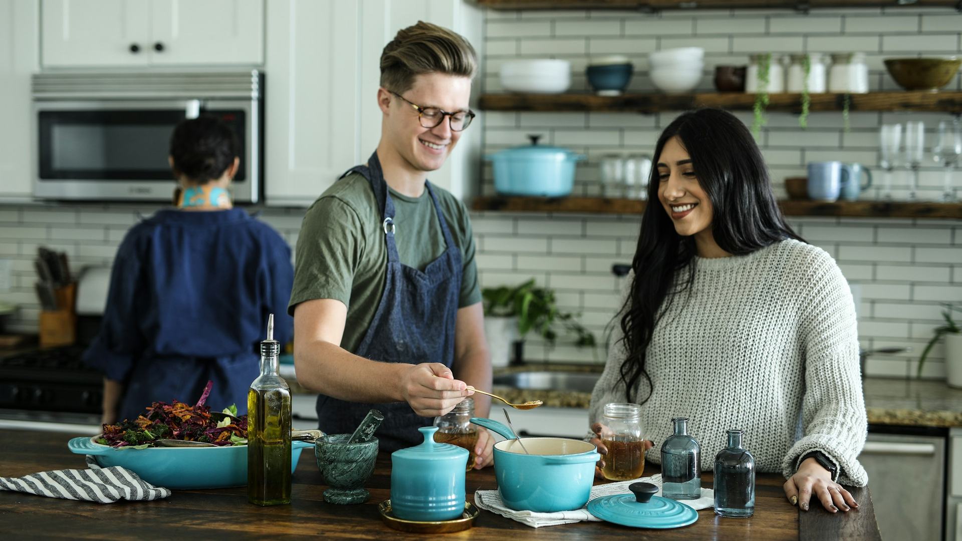 Three people collaborate in a bright modern kitchen with teal cookware, fresh greens, and glass jars on a wooden counter.