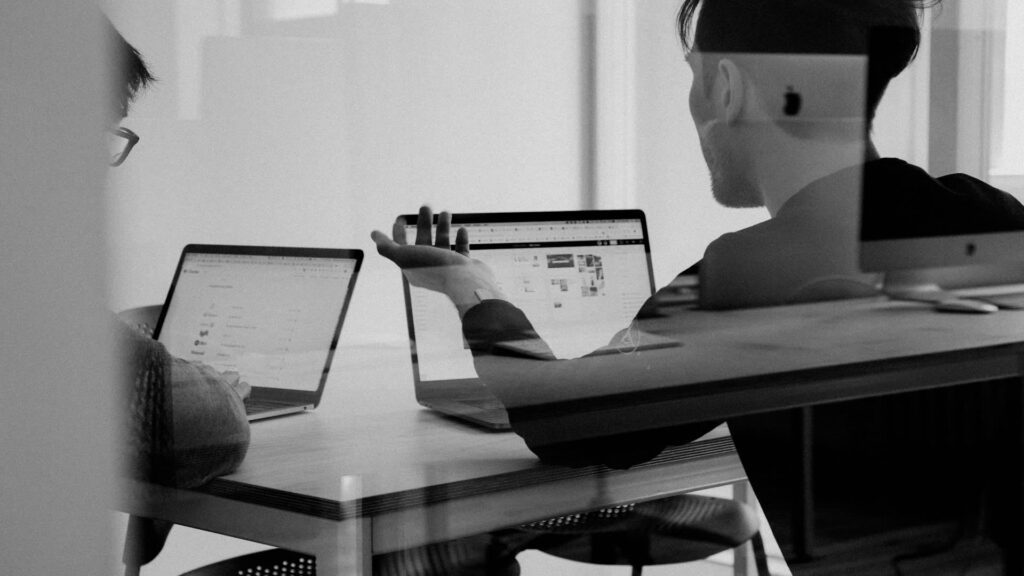 Two IT professionals collaborate over laptops in a modern office, viewed through glass with soft reflections, black-and-white corporate style.