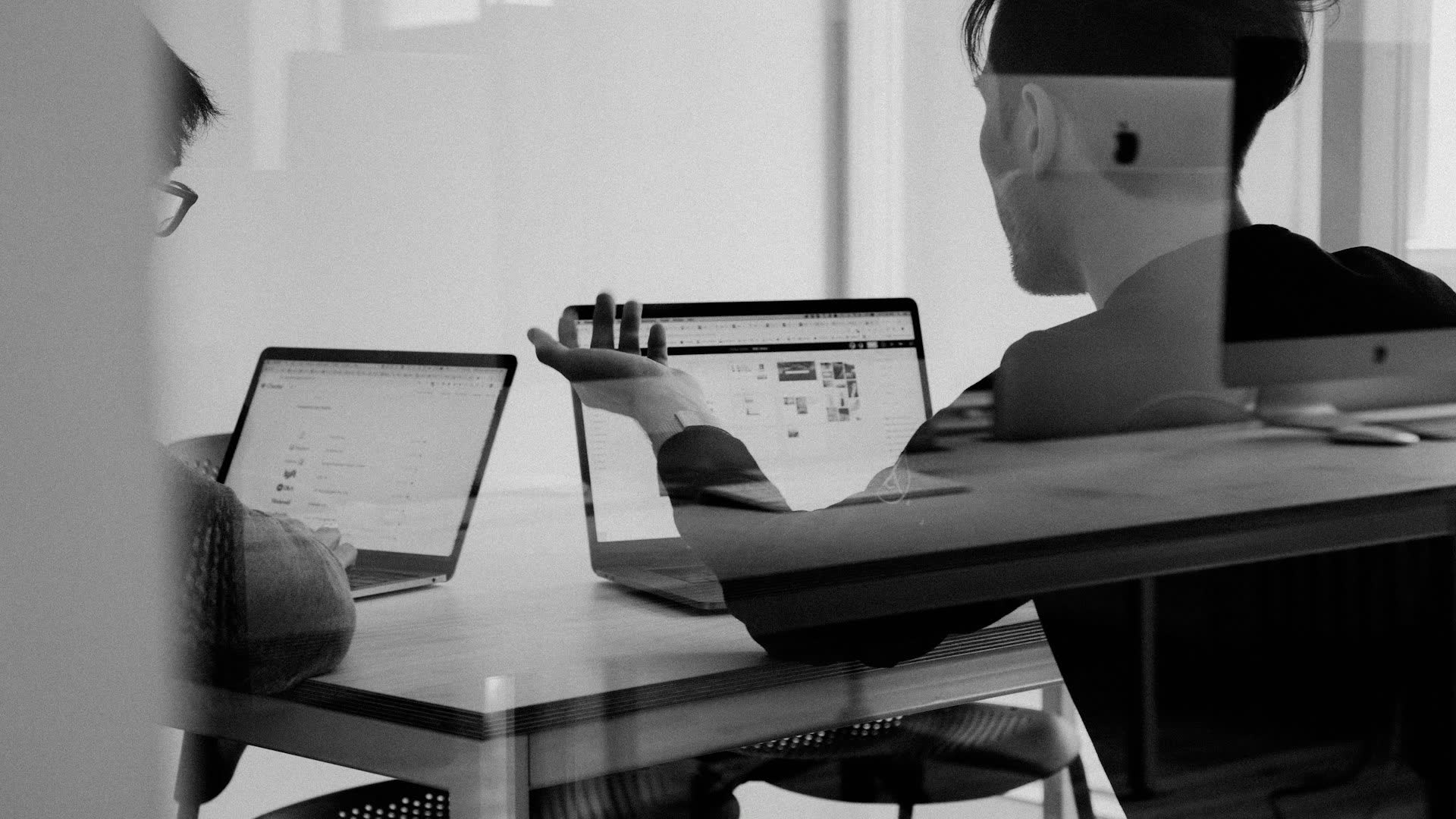 Two IT professionals collaborate over laptops in a modern office, viewed through glass with soft reflections, black-and-white corporate style.