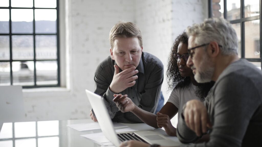 Three colleagues gather around a laptop in a bright office with tall windows, studying the screen with serious, focused expressions