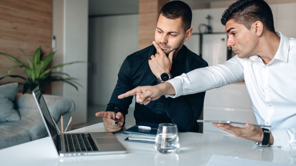 Two colleagues at a white desk study a laptop together, one pointing at the screen while the other thinks it over, during a tense troubleshooting session